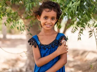 A girl wearing a blue dress looks directly at the camera, holding a bandage up to her recently vaccinated arm. She is smiling. 