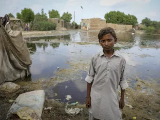 A child looks into the camera, behind him are a flooded field and half submerged homes. 