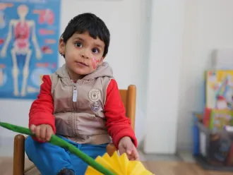 A child with paint on his face, in a classroom-like setting sits on a chair and looks directly into the camera.