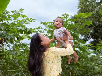 A woman hold up a baby in her arms above her head, behind her are many trees.