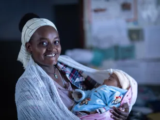 A woman looks at the camera smiling while holding a sleeping baby. 