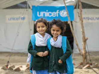 Two young girls wearing blue UNICEF backpacks smile at the camera while standing in front of a UNICEF-funded Temporary Learning Centre.