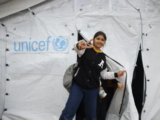 A girl exits a UNICEF tent making the peace gesture with her hand and a bag pack slung over her shoulder.
