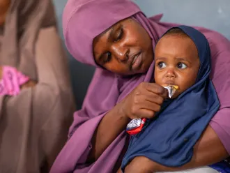 A caring mother in a purple headscarf feeds a young child in a navy hijab, who eats from a snack packet in her arms.