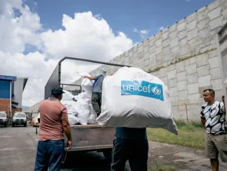 Workers load a large sack with the UNICEF long on it, nto a truck beside a stone wall.