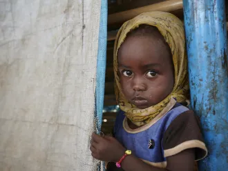 A young child with a serious expression, wearing a yellow headscarf and blue shirt, peeks from behind a curtain