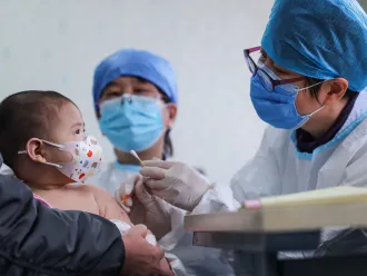 Baby wearing mask looking a doctor in a cap and mask holding a cotton swab with gloved hands