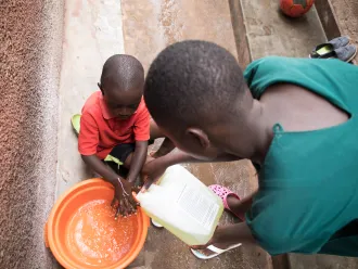 Sharon helps Jonathan to wash his hands with the water and soap mixture at the entrance of their house. In Uganda, the COVID-19 pandemic and lock down has left many families with no option but to stay home and keep stay safe. 
