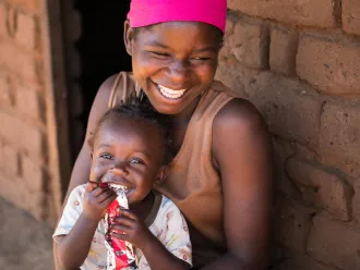 A smiling woman embraces a giggling child holding a packet of peanut paste. 