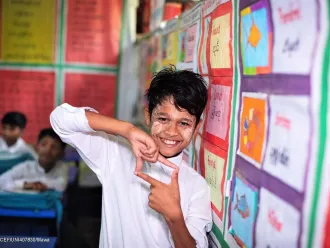A boy uses his hands to create a square photo sign. He is in a classroom setting standing next to bulletin board with many colourful sheets pinned on it. He is wearing a white uniform shirt. In Cox's Bazaar, Bangladesh. 