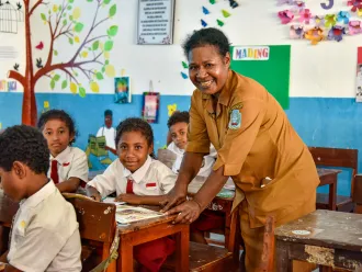 A teacher leans over a students desk and smiles at the camera. The student next to her is also smiling. Photo taken in Indonesia.