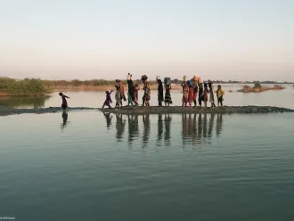 A group of people, including women and children, walk with water containers on their heads along a narrow, muddy path flanked by water in a rural setting.