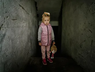 A young girl stands in a dimly lit, narrow concrete passage. She wears a pink polka-dotted vest, holding a stuffed toy, with a somber expression.