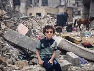 Un enfant assis sur les décombres de la maison familiale regarde l’objectif. On voit derrière lui tous les débris et gravats.