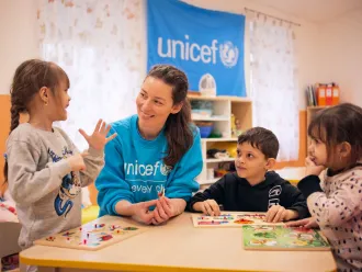 Two girls and a boy play around a table with a woman wearing a blue UNICEF sweater. 