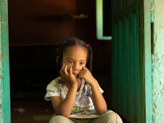 Young girl with pigtails, wearing a patterned shirt, sits pensively in a dimly lit doorway. She rests her chin on her hands, appearing thoughtful.
