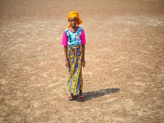 Une jeune fille se tient debout sur une terre aride, portant un chandail rose, une jupe à motifs, un foulard orange et des sandales.