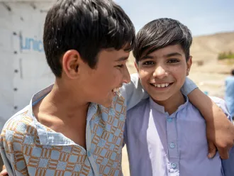 Two adolescent boys standing with their arms around each other’s shoulders and smiling outdoors in the sunshine, in front of a UNICEF tented shelter.