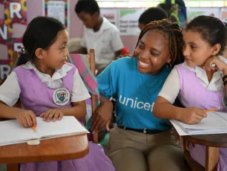 In a classroom, a woman wearing a blue UNICEF shirt crouch and smiles beside two girls sitting at their desk.