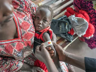 A caregiver holds a young child on her lap. The child wears a red and black outfit as a medical worker measures the child's upper arm with a MUAC tape to assess malnutrition.