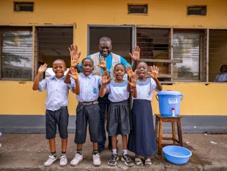 A group of children and a UNICEF worker stand beside a water bucket, smiling at the camera as they show their clean hands.