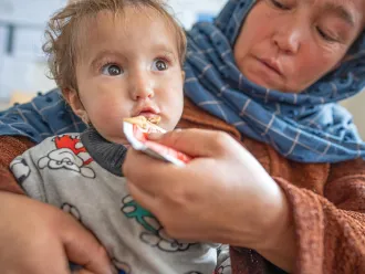 A baby eats a peanut paste out of a packet held by a caregiver in a blue headscarf.