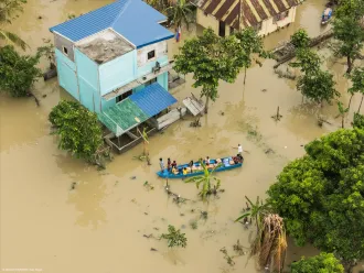  Individuals in a boat traversing a flooded street adjacent to a flooded house, highlighting the effects of severe flooding in the area.