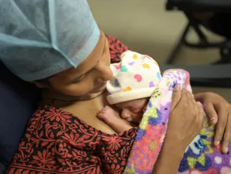A mother wearing a surgical cap lovingly cradles her newborn, wrapped in a vibrant blanket. The baby wears a hat with colorful hearts, exuding warmth and care.