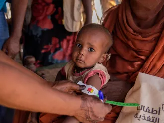 A health worker measures the upper arm of a young child sitting on a woman's lap with a colored band. The setting appears to be an aid center.