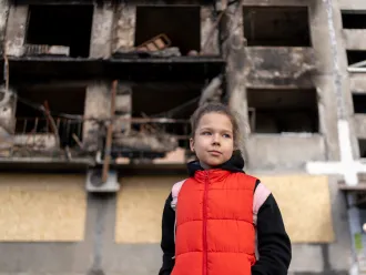Girl wearing a red vest and backpack standing in front of a fire-damaged apartment building.