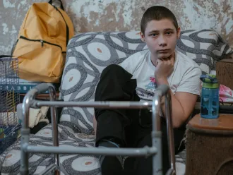 A boy sits on a patterned sofa, looking thoughtful. A walker is in front of him, a yellow backpack and hamster cage are nearby, and a bottle is on a table.