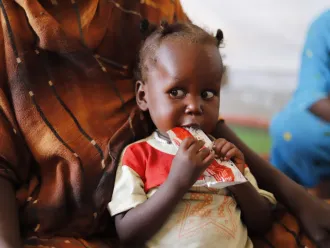 A small child sits on an adult's lap, holding a packet of nutritional paste to their mouth. The child's expression is thoughtful. The adult wears a patterned brown garment.