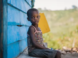 A young child sits on a ledge beside a bright blue wooden wall, gazing thoughtfully at the camera. The background features a blurred, green landscape