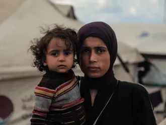 A woman in a purple headscarf and dark clothing holds a young child in a striped sweater. They stand in front of tents, conveying a somber tone.