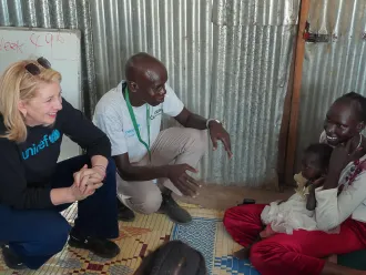 A woman in a UNICEF sweater and a man converse with a smiling woman holding a child, seated on mats in a metal-walled room, conveying warmth and hope.