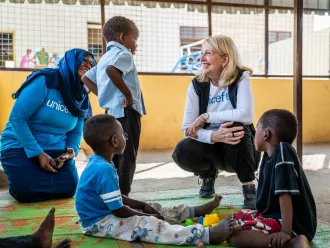 A woman in a UNICEF shirt smiles while kneeling in front of children in a play area. The warm atmosphere is supportive and engaging.