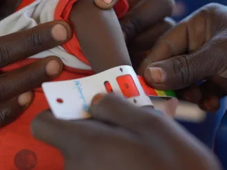 A child's arm is measured with a MUAC tape by two adults, wearing a red and white garment. The scene conveys care and urgency in a healthcare context.