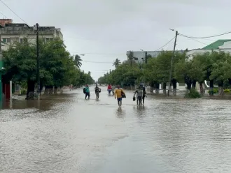 A flooded urban street with submerged road, people wading through knee-deep water, overcast skies, and trees lining the street, conveying a somber mood.