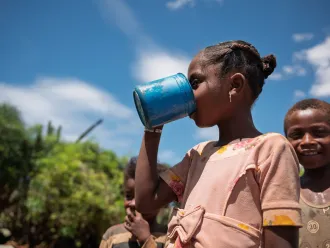 Girl drinking water from a metal cup, with other children nearby outdoors.