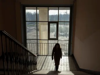 A person walks up a dimly lit staircase, silhouetted against a large window that reveals an overcast outdoor scene. The mood is quiet and contemplative.