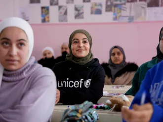 In a classroom with pink walls, a smiling girl wearing a hijab and a UNICEF shirt sits among attentive peers. The atmosphere is warm and positive.