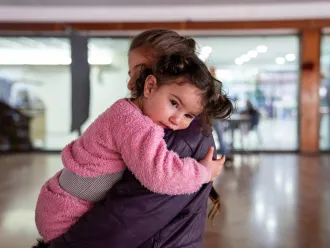 A mother holds a toddler wearing pink clothes in her arms in a brightly lit room. 