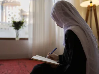 A girl in a white hijab is writing in a notebook, seated on a carpet. 