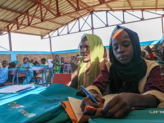 Girls in headscarves writing in notebooks at desks in an open-air classroom.
