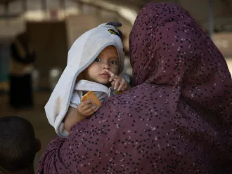 A child with a towel hood is held by a person in a patterned cloak, gazing intently and holding a piece of bread. 