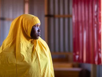 A girl wearing a yellow uniform with a yellow scarf stands by a door looking outside contemplatively. 