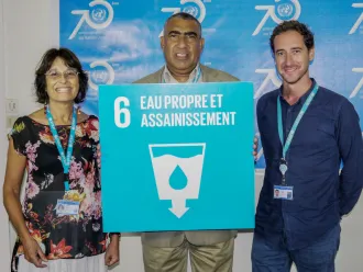 Three people smiling, standing in front of a blue banner, holding a sign that reads "Eau Propre et Assainissement" with the SDG 6 sign that has a water symbol.