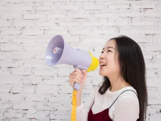 A young girl, standing alone in front of a white wall, smiles while holding up a megaphone.