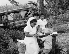 Black-and-white photo of nurses providing a child vaccination beside a jeep in a tropical field; the public-health outreach feels calm and caring.