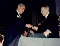 Two elderly men in formal suits exchange a document and handshake at a ceremonial table, with officials seated behind them and flowers nearby.